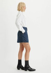 Chemise blanche à manches longues avec détails en volants, associée à une mini-jupe en denim foncé et des bottines noires à bout carré.