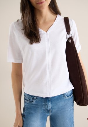 Femme portant un t-shirt blanc à col en V et manches courtes, un jean bleu, portant un grand sac bandoulière en daim brun foncé sur son épaule droite.
