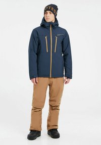 Chaqueta aislante azul marino con detalles en color beige, que cuenta con dos bolsillos en el pecho con cremallera y capucha. Pantalones marrones impermeables y botas negras.