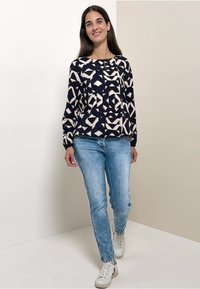 Donna che cammina dentro casa, indossando un top a maniche lunghe blu navy e bianco con motivo geometrico, jeans azzurri e sneakers bianche.