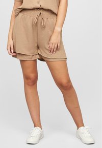 Shorts beiges en tissu texturé, avec une taille élastique et un cordon de serrage, et une coupe ample. Portés avec des baskets blanches.