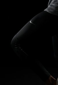 Leggings neri Nike con un design aderente, caratterizzati da un sottile logo argento sulla coscia e linee testurizzate a contrasto lungo i lati.