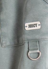 Közeli kép egy szürke szövetről, rajta egy fémcímke "JUICY" felirattal, és egy varrott hurok, amely egy ezüst D-karikát tart.
