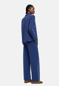 Niebieski zestaw odzieży domowej w dopasowanym stylu, wykonany z miękkiego materiału; składa się z luźnej koszuli z kołnierzykiem oraz spodni o szerokich nogawkach; noszony z brązowymi butami.