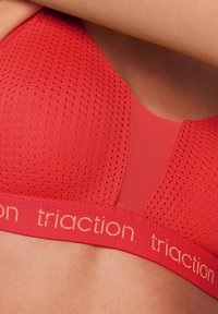 Rød sports-BH med et teksturert, perforert design. Har en glatt band med "traction" merkevare i gull. Lett og pustende materiale.