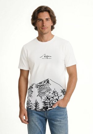 Muž oblečený v bielom tričku s textom „Aspen Where the Wild Begins“ a čiernou skicou hory a stromu, v kombinácii s modrými džínsami.