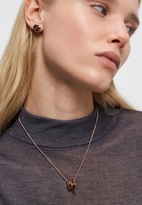 Kvinde iført rosaguld snoede hoop-øreringe og matchende vedhængs halskæde over en mørkegrå højhalset bluse.