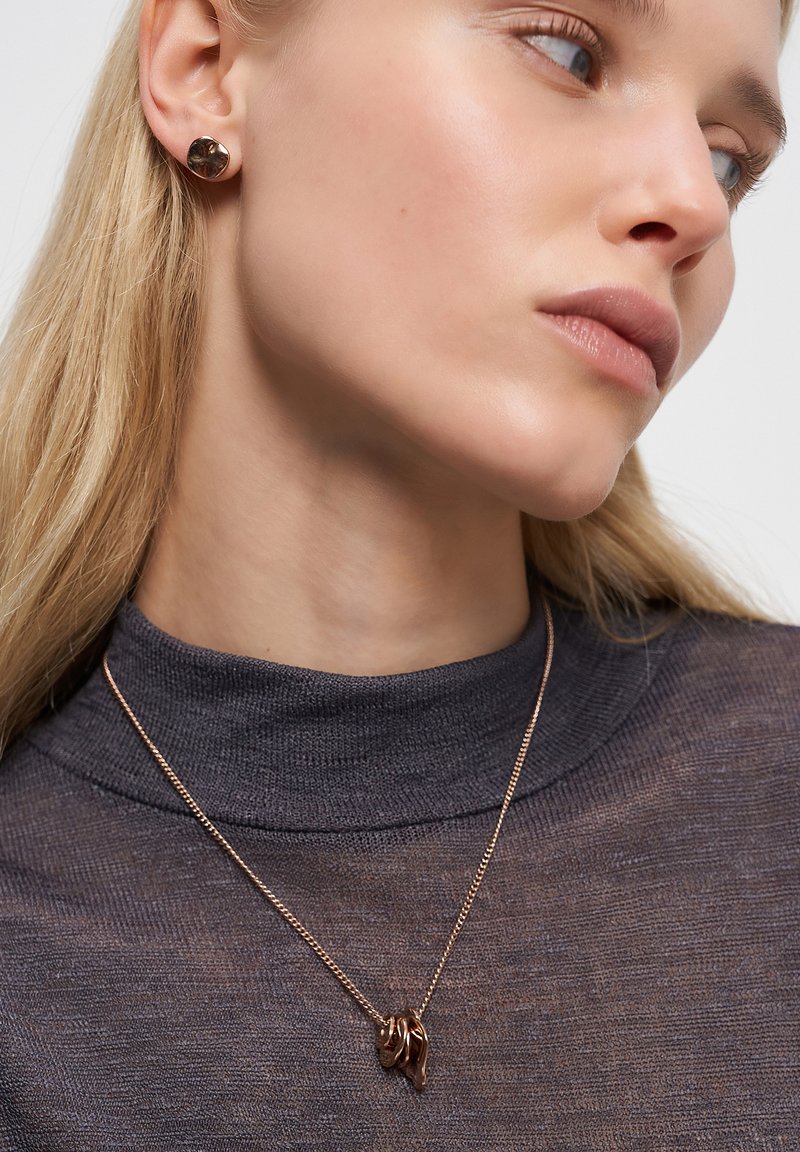 Kvinde iført rosaguld snoede hoop-øreringe og matchende vedhængs halskæde over en mørkegrå højhalset bluse.