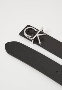 Černý kožený pásek s raženým vzorem a sponou se stříbrným logem "CK." Spona je umístěna ve středu, což zdůrazňuje její elegantní design.