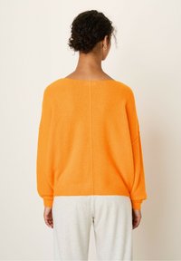 Overdimensioneret, lys orange sweater med en blød tekstur, bred halsudskæring og dropped ærmer. Bageste søm ned langs midten; parret med lysegrå bukser.