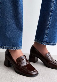 Brązowe lakierowane loafersy z niskim kwadratowym obcasem, z okrągłym noskiem i dekoracyjnym paskiem, w połączeniu z niebieskimi spodniami z szerokimi nogawkami.