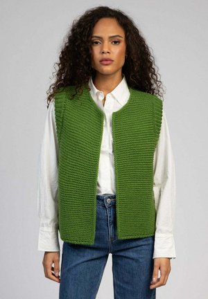 Veste en tricot verte avec un motif texturé, ouverture sur le devant sans fermetures, portée avec une chemise blanche et un jean bleu.