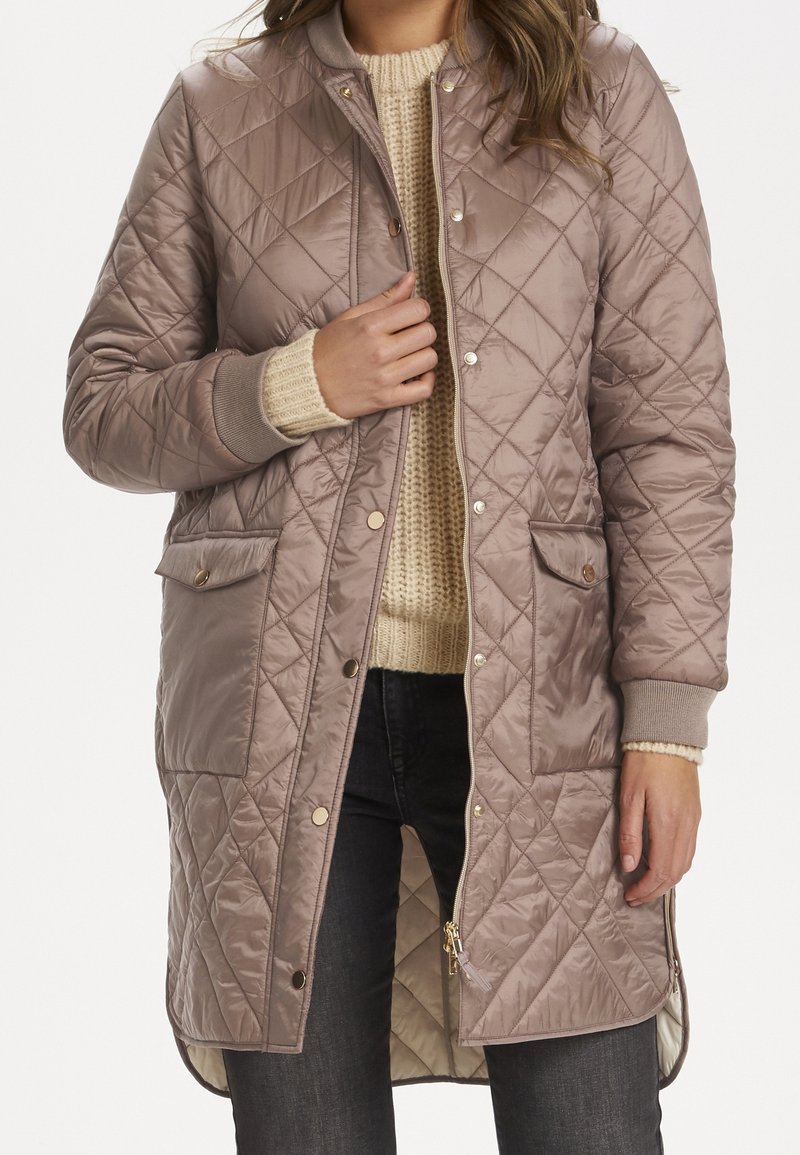 Žena v dlouhém mauve prošívaném kabátu s patentními knoflíky a zipem, pod kterým má krémový pletený svetr a tmavé džíny.