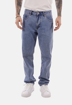 Uomo che indossa jeans chiari a vestibilità comoda, sneakers bianche, t-shirt bianca e braccialetto nero con perline, in piedi con le mani rilassate.