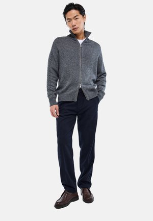 Homme portant un pull zippé gris avec des manches texturées, un pantalon bleu marine et des chaussures en cuir marron, debout devant un fond clair uni.