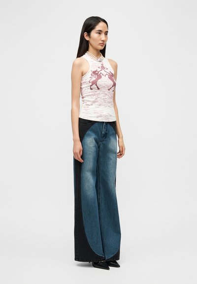 Λευκό αμάνικο τοπ με ροζ γραφικό σχέδιο, συνδυασμένο με παντελόνι τζιν με φαρδιά μπάλα, με μπλε και μαύρα χρωματιστά μπλοκ. Μαύρα μυτερά παπούτσια.