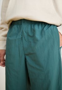 Persoon in teal elastische taillebandbroek en crèmekleurige sweatshirt, met één hand in de zak en de andere ontspannen naast het lichaam.
