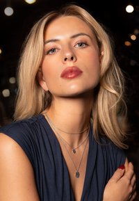 Des colliers superposés en argent avec divers designs de pendentifs, portés sur un haut en tissu bleu foncé. De douces cheveux blonds encadrent le visage.