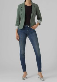 Blazer vert avec revers, manches longues et poignets retroussés, associé à un jean skinny bleu foncé et des escarpins argentés.