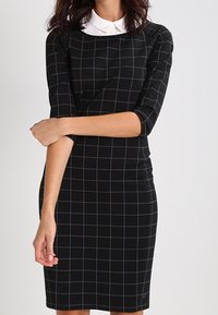 Nainen, jolla on päällään musta istuva mekko, jossa on valkoinen ruudukko ja valkoinen kaulus, kädet ristissä vyötäröllä tasaisen taustan edessä.