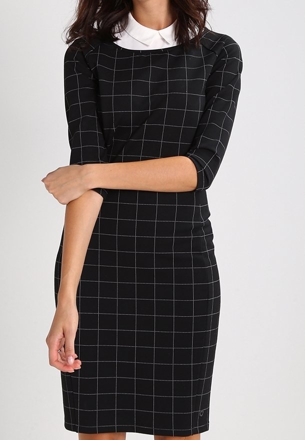 Nainen, jolla on päällään musta istuva mekko, jossa on valkoinen ruudukko ja valkoinen kaulus, kädet ristissä vyötäröllä tasaisen taustan edessä.