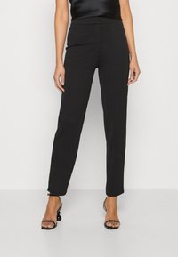 Pantalon noir sur mesure à coupe droite, confectionné dans un tissu lisse. Associé à des sandales à talons fines noires.