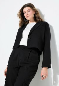 Blazer cropped nero con chiusura a un bottone, rever a punta e maniche lunghe. Indossato sopra una t-shirt bianca con testo. Tessuto testurizzato.