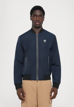 Navy bomber jacket with a zip front and ribbed cuffs. Features two side pockets and a textured logo on the chest. Smooth fabric finish.