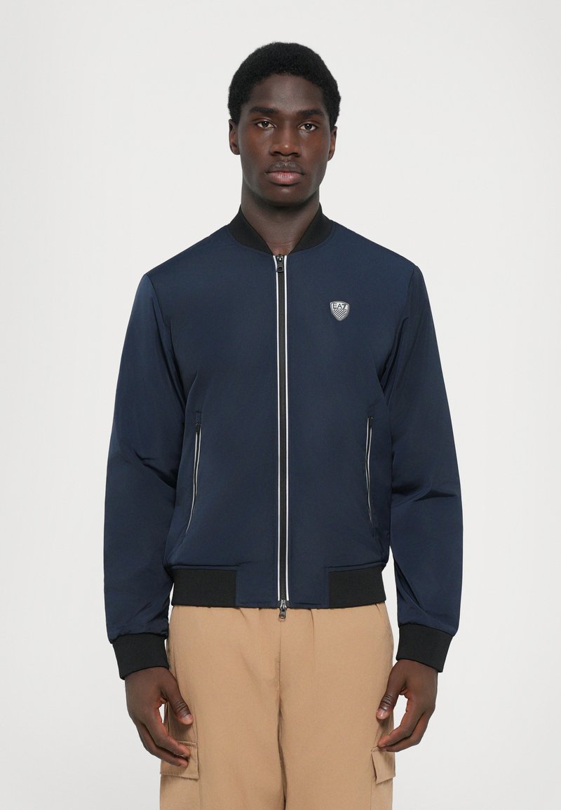 Casaco bomber azul-escuro com fecho zip e punhos canelados. Apresenta dois bolsos laterais e um logotipo texturizado no peito. Acabamento de tecido liso.