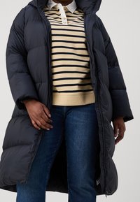 Person trägt eine lange schwarze Steppjacke über einem beige- und schwarzgestreiften Hemd mit Kragen und goldenen Knöpfen sowie einer blauen Jeans.