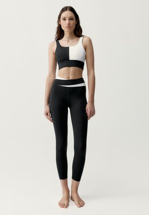 Junge Frau steht barfuß und trägt einen schwarz-weißen Sport-BH im Color-Blocking-Stil sowie hoch taillierte Leggings vor einem schlichten hellen Hintergrund.