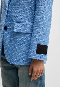 Blauer strukturierter Blazer mit einem markanten Bobble-Muster, Einzelknopfverschluss und einem schwarzen Etikett mit "HUGO" auf dem Ärmel.