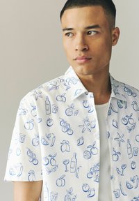 Chemise à manches courtes en tissu blanc ornée d'un imprimé bleu de fruits et de boissons, avec un col étalé et une fermeture à boutons.