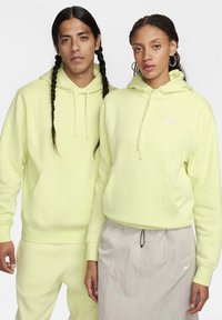 Uomo e donna che indossano felpe Nike giallo chiaro abbinate, l'uomo con pantaloni abbinati, la donna con una gonna grigia chiara con il logo Nike.