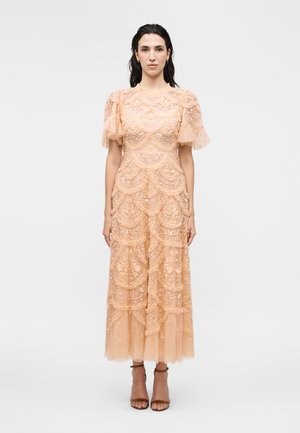 Kvinde iført en ferskenfarvet, ankel-lang blondekjole med puffede korte ærmer og blomsterbroderi, kombineret med brune ankelstroppesko.