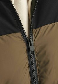Veste à zip olive et noire avec une finition mate, des panneaux en tissu contrastés et une fermeture éclair noire robuste avec détails de la marque.