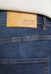 Denim jeans with a dark blue wash, featuring a leather patch on the back with "THE DENIM JOURNEY" embossed, and contrast stitching details.