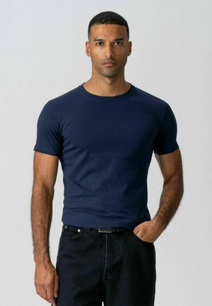 Mand iført tætsiddende marineblå kortærmet t-shirt og sorte jeans med den ene hånd i lommen, stående mod en ensfarvet lys baggrund.