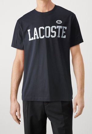 Mężczyzna w czarnym t-shircie z krótkim rękawem z dużym białym napisem "LACOSTE" i małym owalnym logo z krokodylem na piersi.