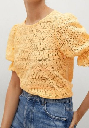 Vrouw die een gele kanten blouse met pofmouwen draagt, gestopt in een hoge taille blauwe spijkerbroek, met één hand op haar heup.