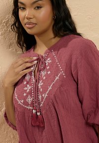 Bordeaux tuniek van gestructureerde stof, met witte borduursels en kwastjes bij de halslijn. Losse pasvorm met driekwart mouwen.