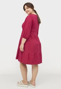 Rotes knielanges Kleid mit dreiviertellangen Puffärmeln, gestuftem Design und weicher Textur. Kombiniert mit hellen Sandalen.