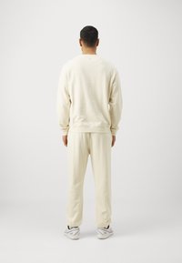 Krämfärgad sweatshirt och matchande byxor i mjukt tyg. Designen har ribbade muddar och en avslappnad passform. Fötterna har vita skor.