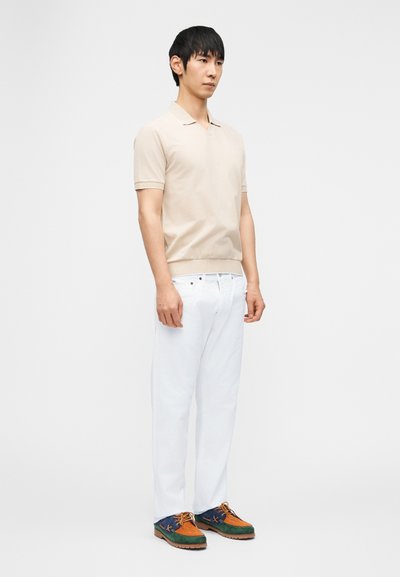 Άνδρας που στέκεται φορώντας μπεζ κοντομάνικο πόλο πουκάμισο, λευκό παντελόνι και πολύχρωμα καφέ, πράσινα και ναυτικά δετά παπούτσια σε λευκό φόντο.