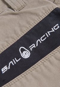 Tissu beige avec un patch horizontal noir brodé du texte blanc "Sail Racing" et un logo circulaire ressemblant à une cible au centre.