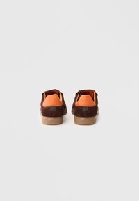 Ténis em camurça castanha com uma aba do calcanhar laranja e sola de borracha texturizada castanha. Apresenta um design minimalista e detalhes de costura subtil.
