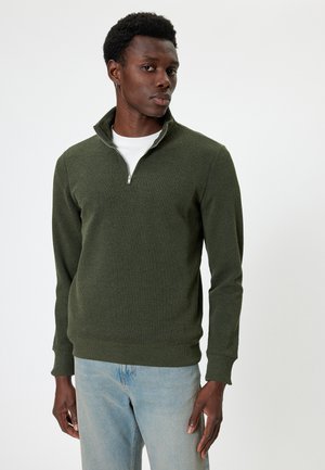 Groene geribbelde trui met een ritskraag, gecombineerd met lichtblauwe jeans. De stof lijkt textuur te hebben, met een getailleerde pasvorm.