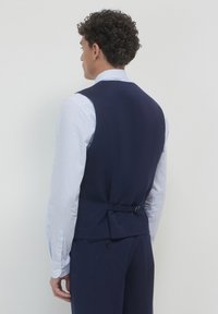 Uomo con capelli ricci che indossa un gilet e pantaloni a righe blu navy, abbinati a una camicia a righe bianche e blu, visto da dietro.