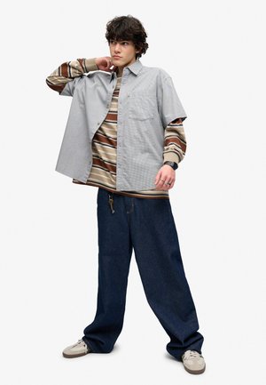Ung mand med krøllet hår iført stribet langærmet skjorte under kortærmet knap-skjorte, løsthængende mørke jeans og sneakers, poserer med hånd på halsen.