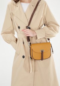 Gelbe Leder-Umhängetasche mit braunem Riemen und silberfarbenen Beschlägen. Die Tasche hat einen Klappenverschluss und eine strukturierte Oberfläche.